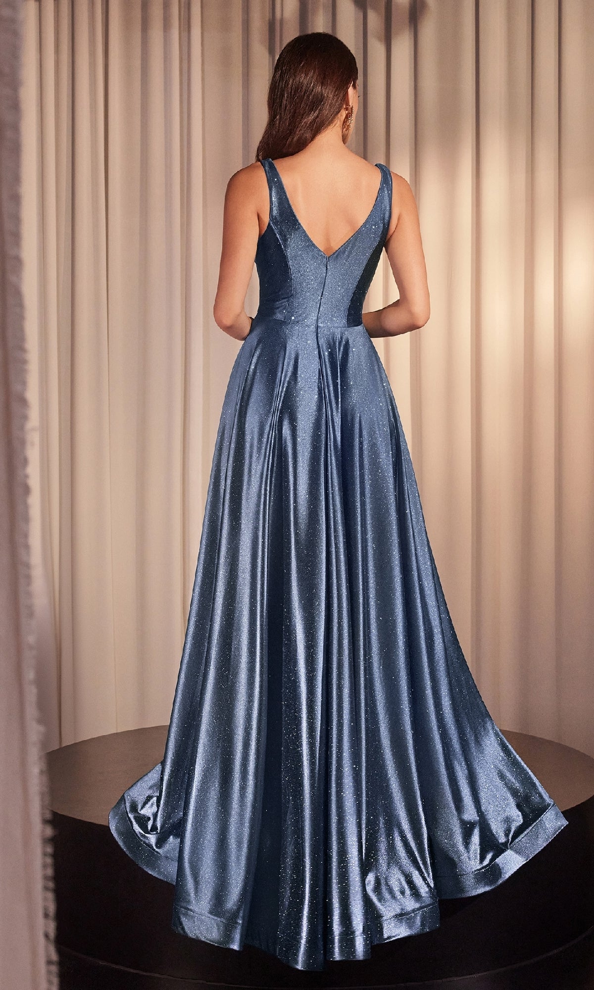 A woman stands on a platform with her back to the camera, wearing the sleeveless, floor-length, blue V-Neck Long Glitter Prom Dress Ladivine CC2349. Cream-colored curtains hang in the background.